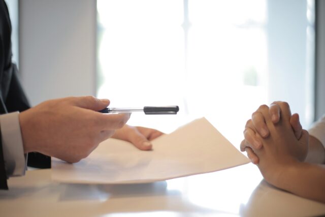 Two people sit across desk, with pen and paper in one's hand, implying negotiations or conflict resolution are being discussed