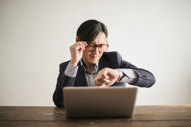 Employee at computer looking at watch representing Scheduling conflicts in the workplace