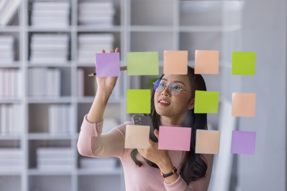 Business female employee with many conflicting priorities arranging sticky notes commenting and brainstorming on work priorities colleague in a modern office.