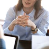 Smiling woman attentively listening to partners concentrated on business negotiations, nonverbal communication concept, focus on locked crossed fingers, clenched hands gesture close up view