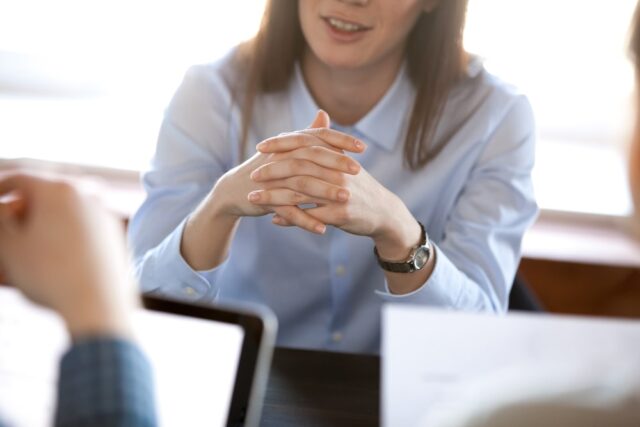 Smiling woman attentively listening to partners concentrated on business negotiations, nonverbal communication concept, focus on locked crossed fingers, clenched hands gesture close up view