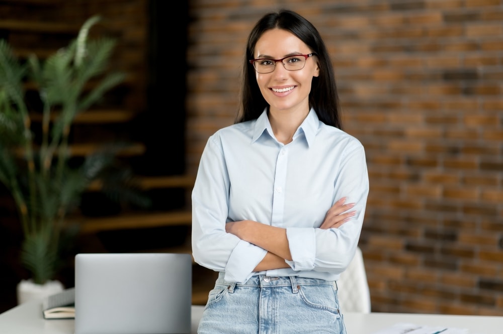 Portrait of successful confident pretty young adult caucasian businesswoman or broker, stand near work desk in office, wearing formal stylish clothes, arms crossed, looking at camera, smiles friendly