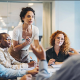 A diverse group of office professionals engaged in a serious meeting. A woman stands at the table leading the discussion with a focused expression, while colleagues listen intently and take notes, illustrating a difficult but productive workplace conversation.
