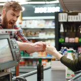 A high-tension scene at a grocery checkout where an aggressive male customer in a plaid shirt is shouting at a startled female cashier. A bottle of olive oil and a computer monitor are on the counter.